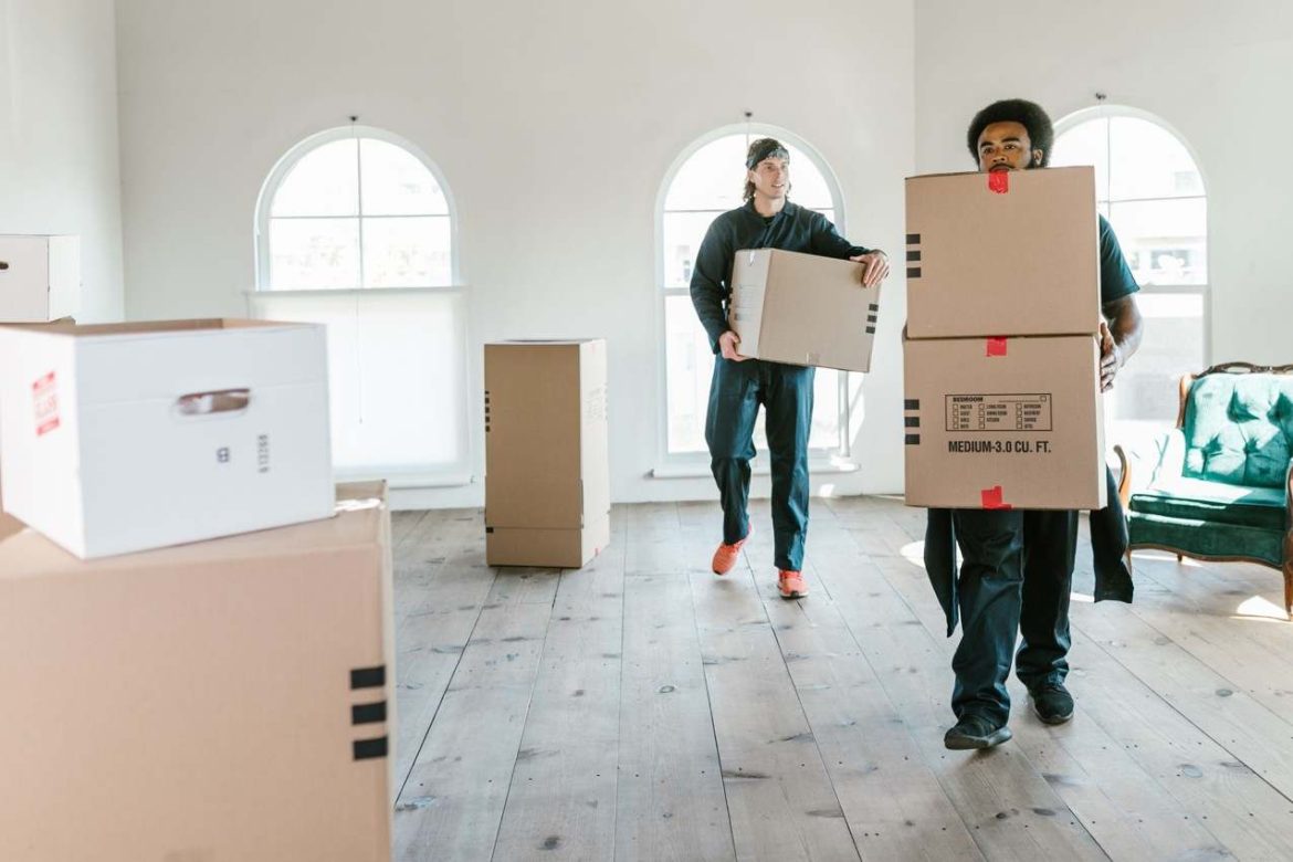Deux déménageurs professionnels transportent des cartons dans un appartement lumineux au sol en bois, illustrant la question : est-il plus rentable de déménager seul ou avec un professionnel.