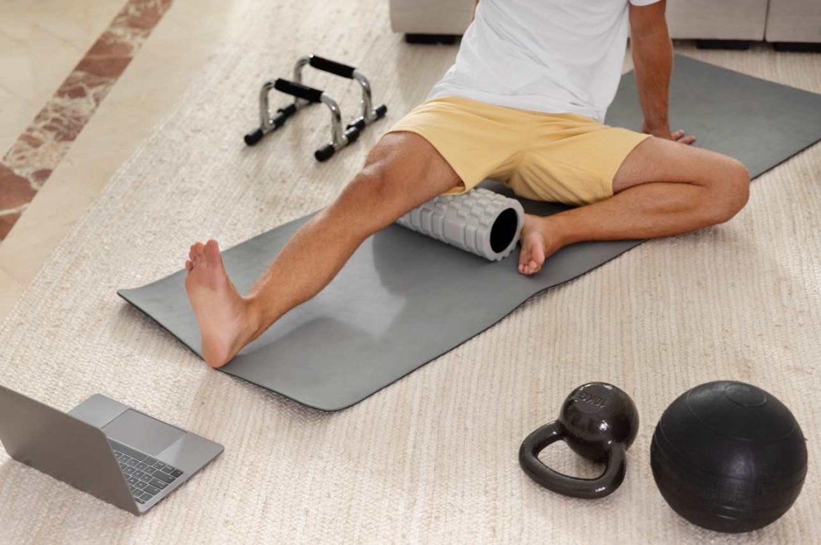 Homme assis sur un tapis de fitness effectuant un automassage des cuisses avec un rouleau de massage grisé à picots.