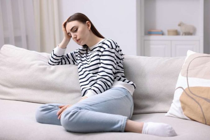 Une jeune femme assise sur un canapé, la main sur le front, cherchant des solutions pour surmonter la fatigue chronique.